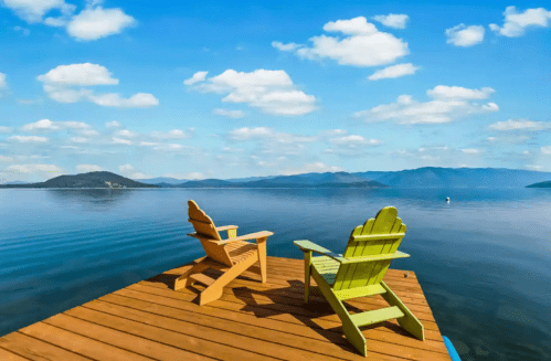 Two colorful chairs on a wooden dock overlooking a calm lake and distant mountains under a blue sky with fluffy clouds.